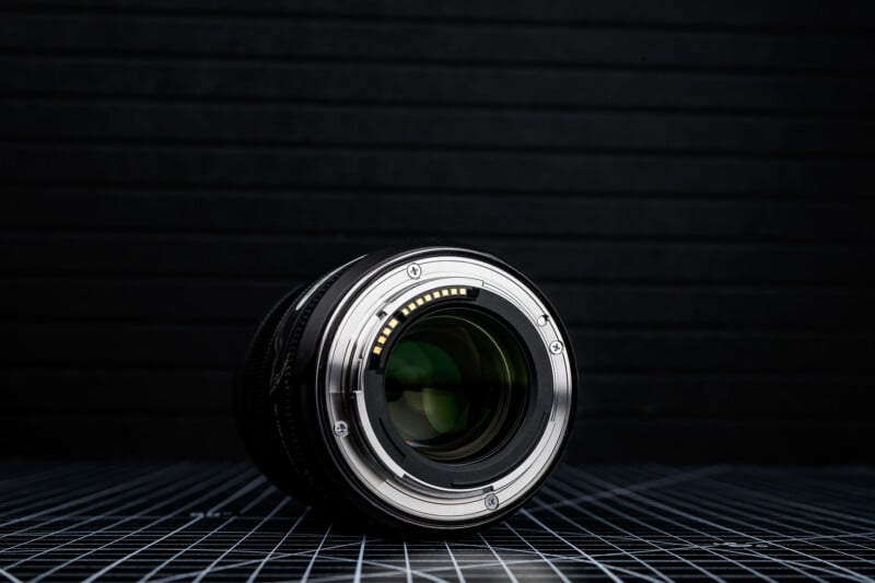 A close-up of a camera lens lying on its side, showing the metal mount and electronic contacts, with a dark, grid-patterned background.