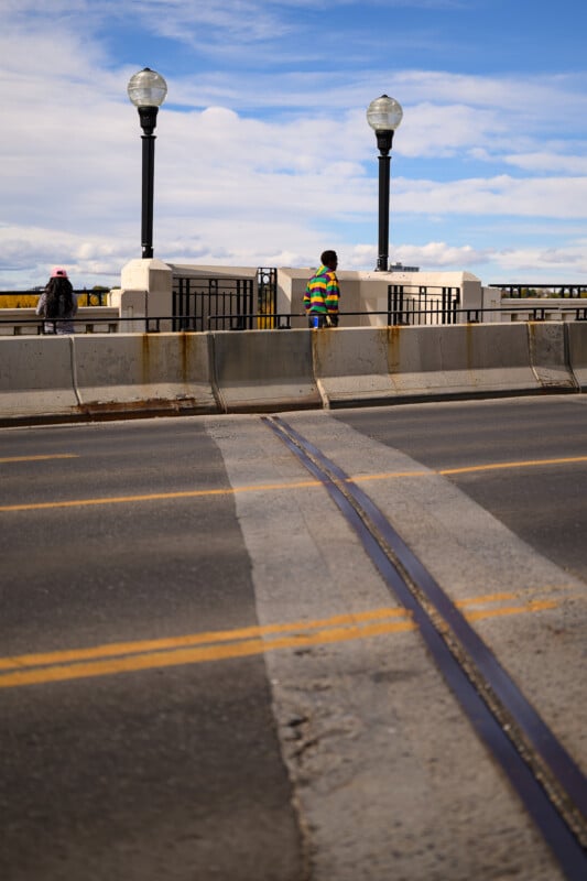 A person in a colorful striped sweater stands by a fence between two street lamps on a bridge, while another person walks away to the left. The sky is partly cloudy and tracks cross the road in the foreground.
