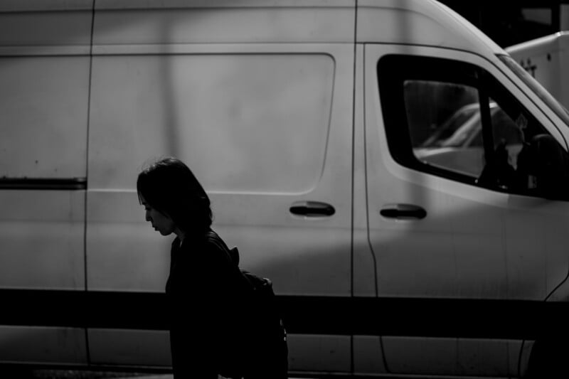 A person in dark clothing walks in front of a white van, their face partially in shadow. The image is in black and white, with strong contrasts and a somber atmosphere.