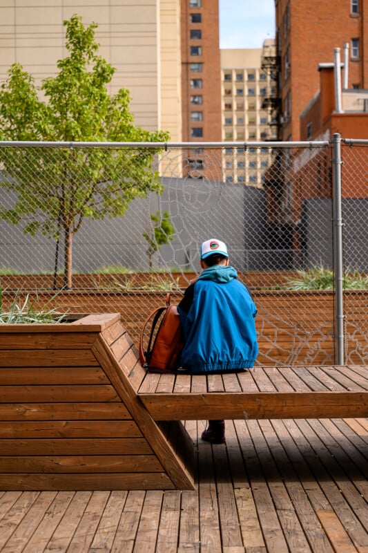A person in a blue jacket and white cap sits alone on a wooden bench in an urban outdoor area, facing away. There are buildings, a chain-link fence, and a small green tree in the background.
