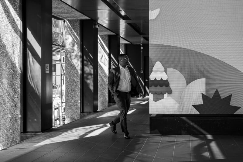 A man in a suit walks through a modern building corridor, partially lit by sunlight. He passes by a large, colorful graphic display featuring abstract shapes. The image is in black and white.