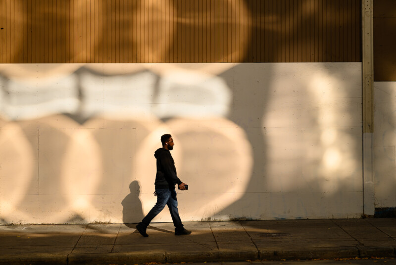 A man in a dark hoodie and jeans walks along a sidewalk in sunlight, casting a shadow against a wall with large, circular light patterns.