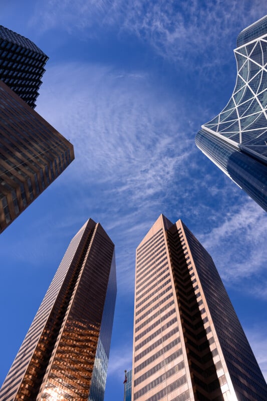 Tall skyscrapers with reflective glass facades rise into a blue sky with wispy clouds, viewed from below, creating a dramatic and modern urban cityscape.