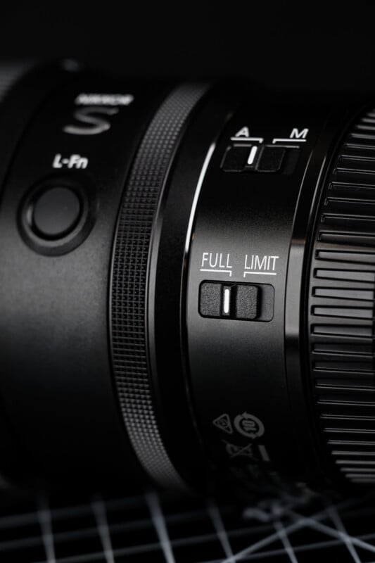 Close-up of a black camera lens showing switches labeled "A M," "FULL LIMIT," and "L-Fn" with white markings, and a textured focus ring on the right side. The background is dark and slightly out of focus.