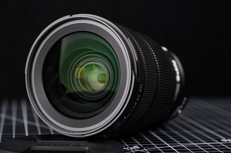 A close-up of a camera lens with a black body and green reflections on the glass, resting on a grid-patterned surface. The lens cap is placed nearby, and the background is dark and out of focus.