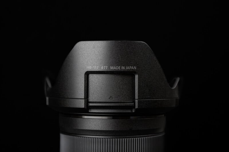 Close-up of a camera lens with a lens hood attached, labeled "HB-117 477 MADE IN JAPAN," set against a black background.
