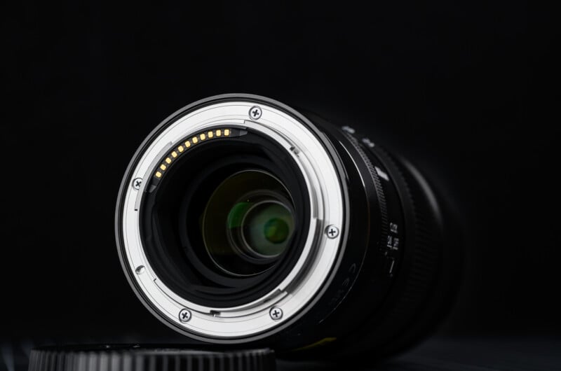 Close-up of the metal mounting side of a camera lens, showing electronic contacts and lens glass, with a black background and part of a lens cap visible at the lower edge.