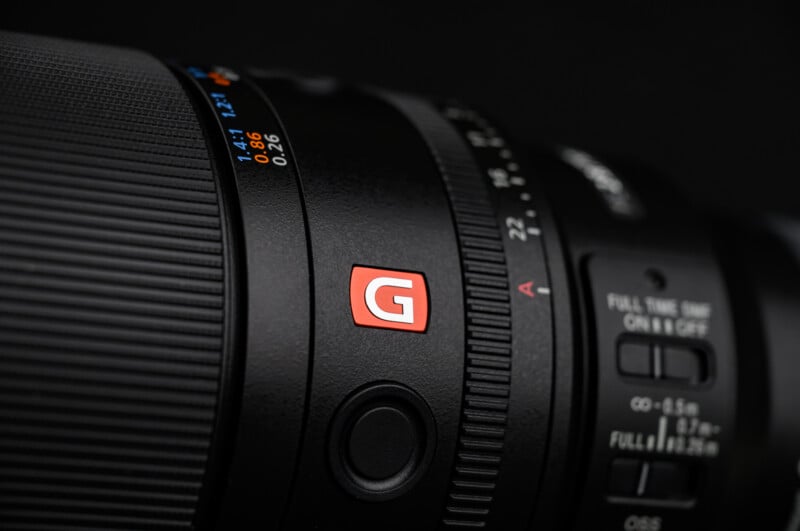 Close-up of a black camera lens featuring a red "G" badge, focus and zoom rings, aperture markings, and several switches, set against a dark background.