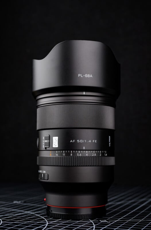 A black camera lens with a lens hood, labeled "PL-68A" and "AF 50/1.4 FE," stands upright on a grid-patterned surface against a dark background.