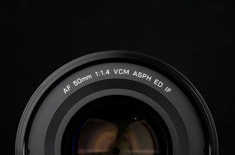 Close-up of a camera lens against a black background, showing the text "AF 50mm 1:1.4 VCM ASPH ED IF" around the front rim.