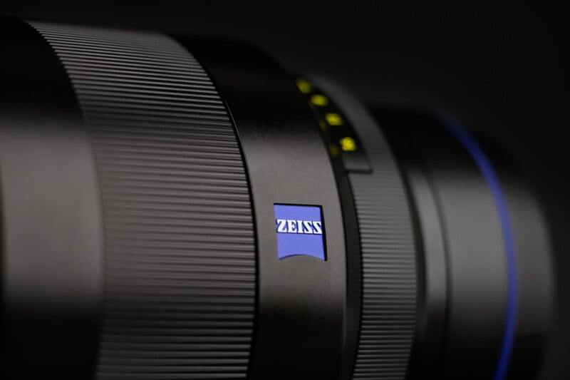 Close-up of a black camera lens featuring the blue and white ZEISS logo, with fine texture and ridges visible on the lens barrel against a dark background.