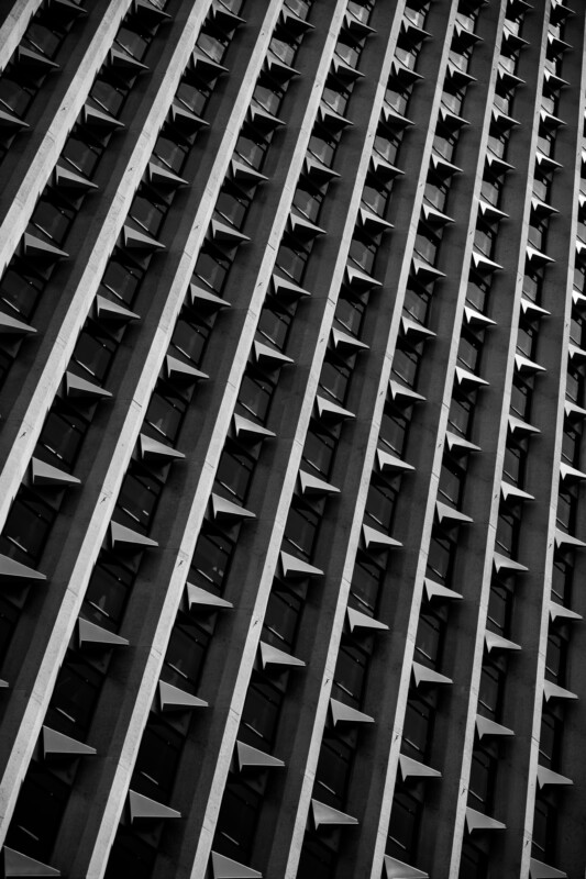 A black and white photo of a tall building façade, viewed at an angle, with repeating vertical columns and rows of small protruding shades beneath each window, creating a geometric, rhythmic pattern.
