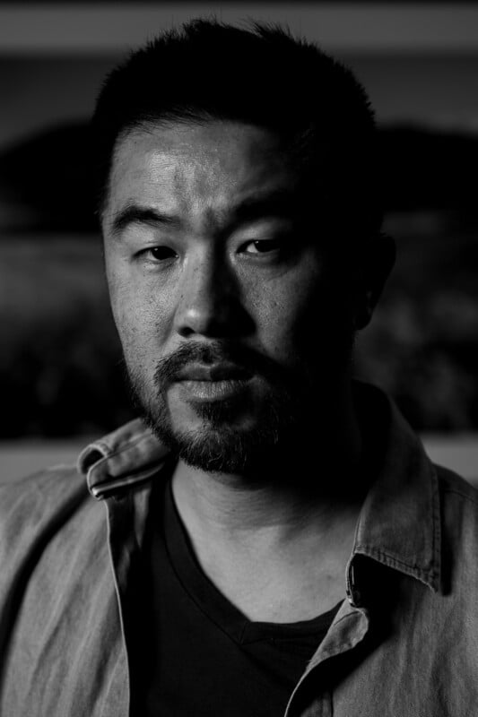 A man with short hair and a beard looks seriously at the camera. The black and white photo has dramatic lighting, casting shadows on half of his face, and he wears a collared shirt over a t-shirt.