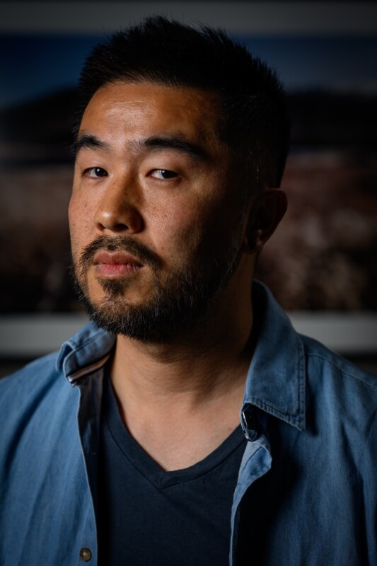A man with short black hair and a trimmed beard poses against a blurred background, wearing a dark t-shirt layered with a light denim shirt. He looks slightly to the side with a neutral expression.
