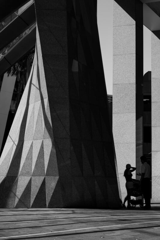 A woman with a stroller and a man stand in shadow beneath large geometric stone columns, with dramatic light and angular shadows creating a striking urban scene.