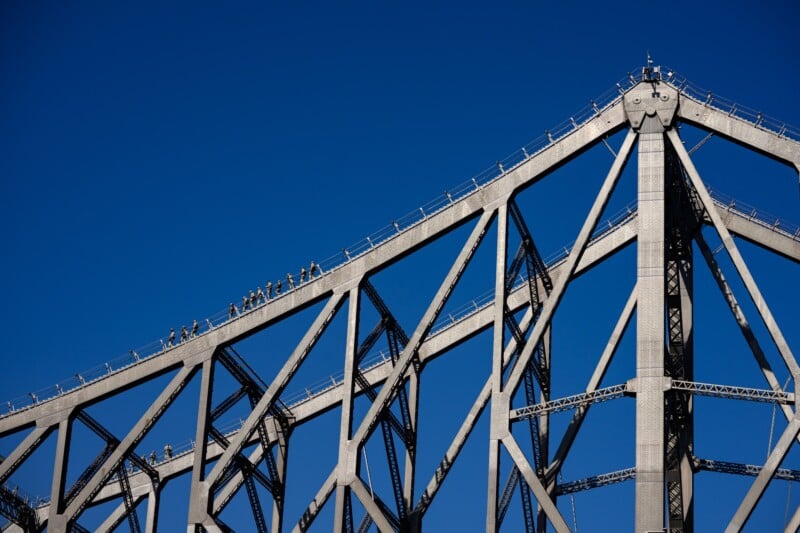 A group of people walk along the top of a large steel bridge structure with beams and railings, set against a clear blue sky.