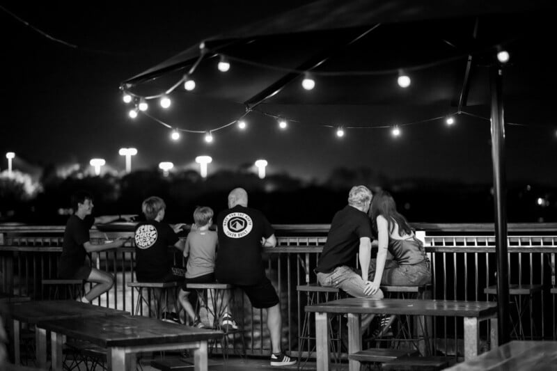A group of people sit on stools at an outdoor patio at night, under string lights. Two couples sit close together and two children sit nearby, looking out over the railing towards a dark, softly-lit landscape.