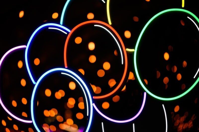 Bright neon circles in blue, red, orange, green, and white overlap against a dark background, with orange bokeh lights scattered throughout, creating an abstract and vibrant composition.