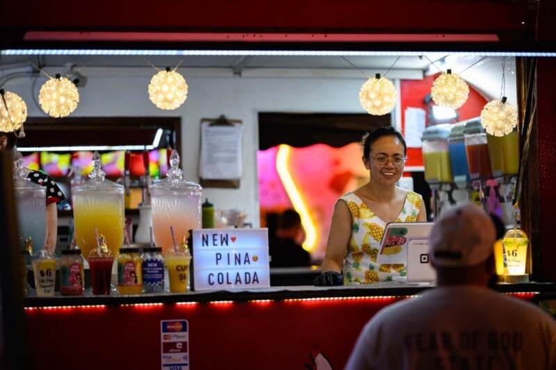 A woman wearing glasses and a pineapple-print dress smiles while serving drinks at a colorful outdoor bar. A sign advertises a new piña colada flavor, and various drinks are displayed in clear dispensers.