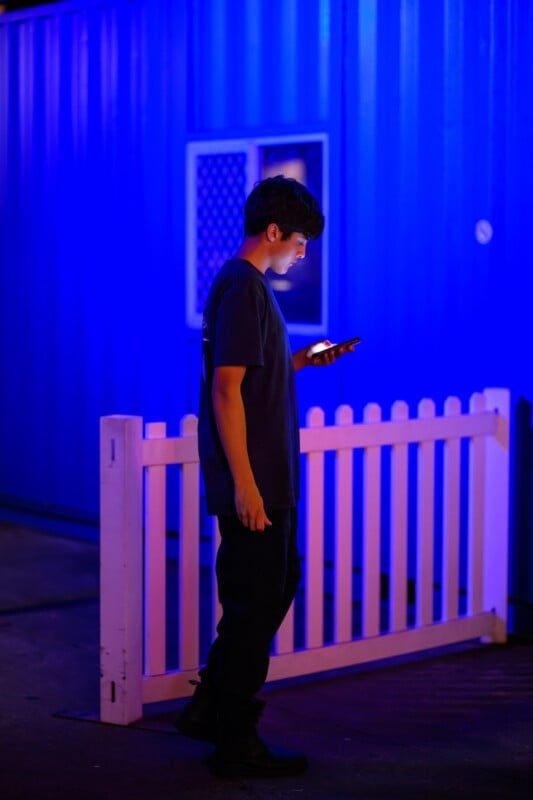A person stands near a white picket fence at night, illuminated by blue and purple lighting, looking at a smartphone. A blue wall and window are in the background.