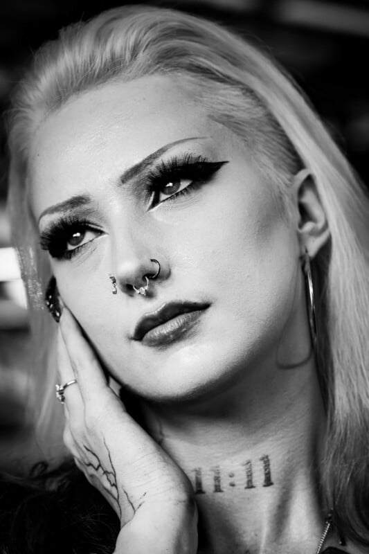 Black and white portrait of a person with platinum blonde hair, dramatic eyeliner, nose piercings, hoop earrings, and a neck tattoo reading "11:11." They are gazing upward with a thoughtful expression, hand resting on their face.