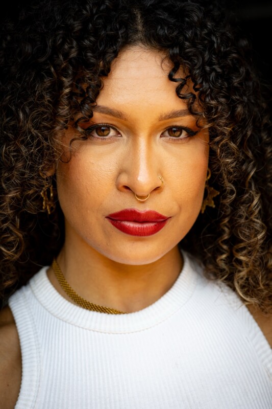 A person with curly brown hair, bold red lipstick, gold hoop nose ring, and gold earrings, wearing a white sleeveless top, looks confidently at the camera against a dark background.