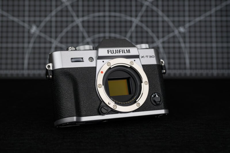 A Fujifilm X-T30 mirrorless camera body with no lens attached, placed on a black surface with a geometric grid backdrop.