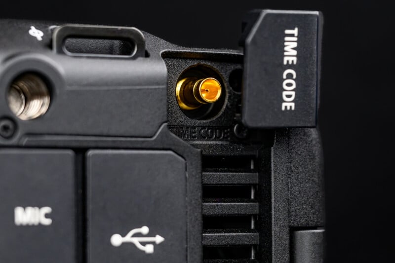 Close-up of a camera or audio device port labeled "TIME CODE," with the protective cover open, revealing a gold connector. Surrounding ports for MIC and USB are partially visible.