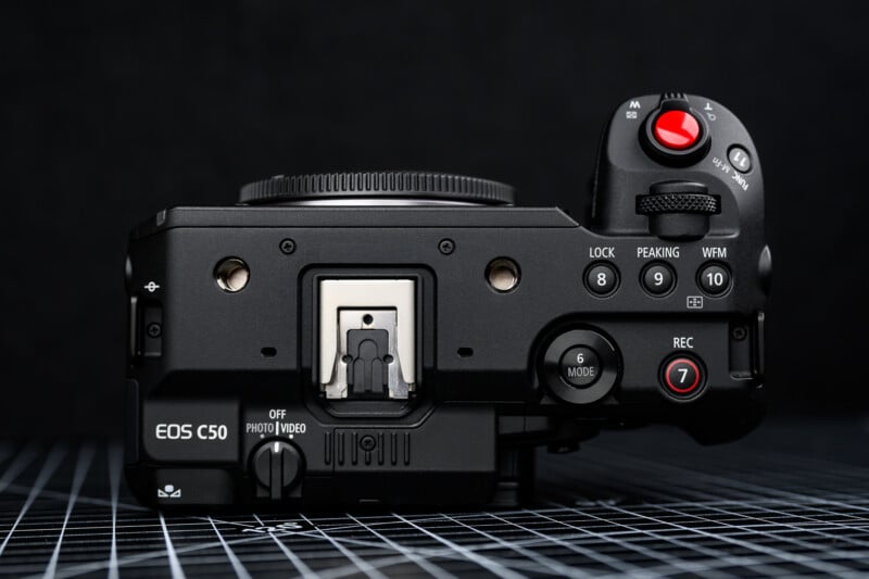 Top view of a Canon EOS C50 camera body on a checkered surface, showing various buttons, dials, and the hot shoe mount, with a black background.
