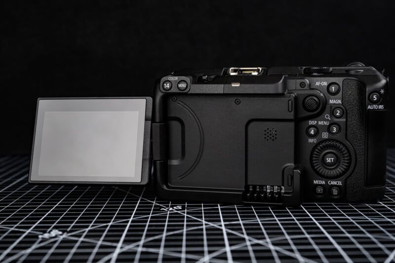 A digital camera with its LCD screen flipped out to the side is placed on a black grid-patterned surface, showing the back view with various buttons and controls visible.