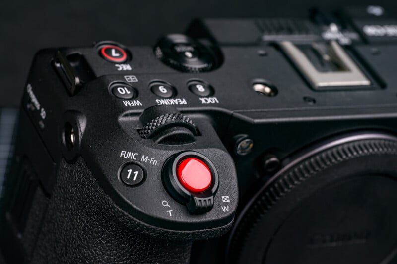 Close-up of a black digital camera showing a red shutter button, various control dials, labeled buttons, and textured grip on the left side of the camera body.