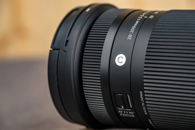 Close-up of a black camera lens, showing the focus and zoom rings, AF/MF switch, and "20-200mm" marking. The background is blurred in beige and gray tones.