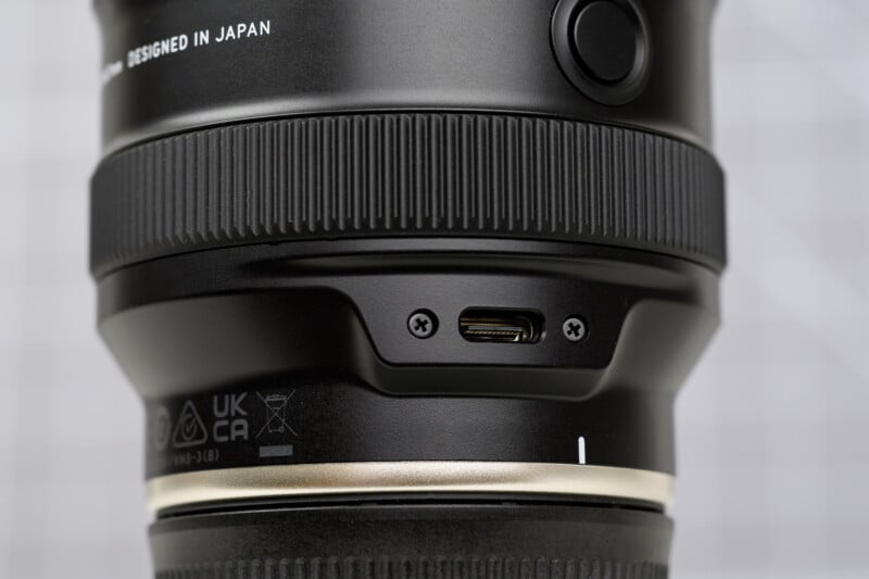 Close-up of a camera lens showing a USB-C port and two screws on its body. The lens has a ridged focus ring, and “DESIGNED IN JAPAN” text is partially visible at the top.