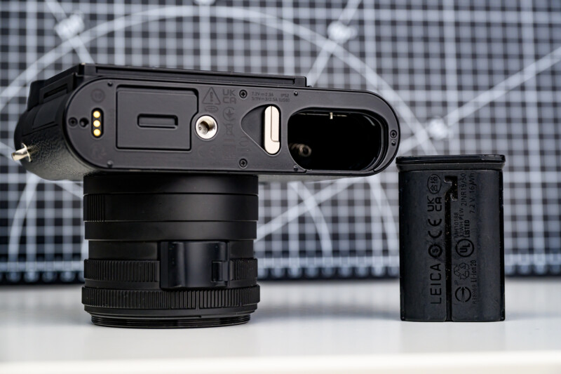 Close-up of a camera lying on its side with the battery compartment open and the battery removed, placed next to the camera. A grid background with white lines is visible behind them.