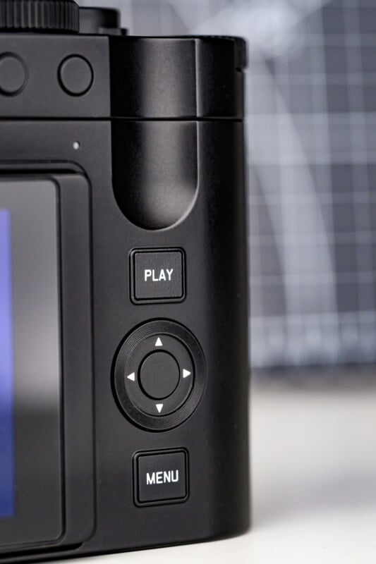 Close-up of a camera’s back panel showing three buttons labeled PLAY, MENU, and a circular directional pad with four arrows, next to part of the screen and a blurred grid background.