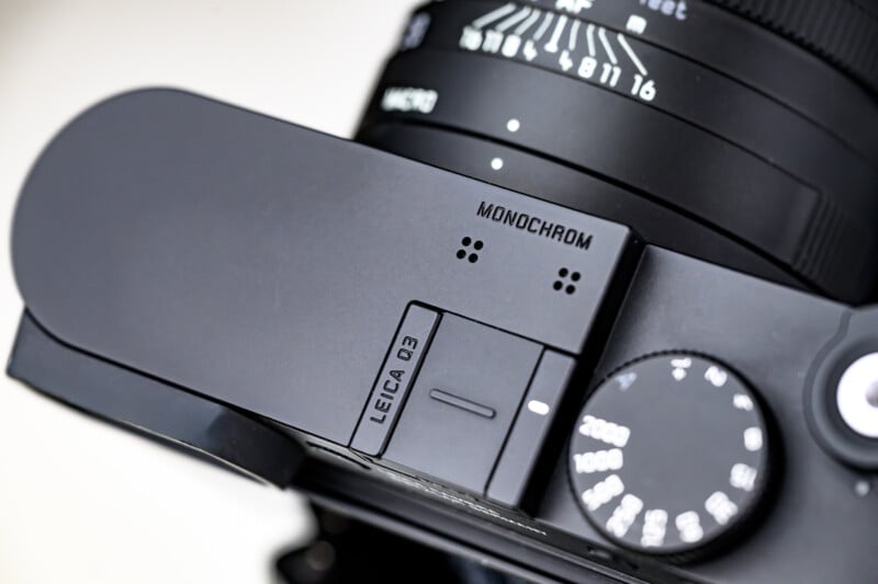 Close-up of the top of a Leica Q3 Monochrom camera, showing the model name, control dials, and part of the lens with aperture and focus markings.