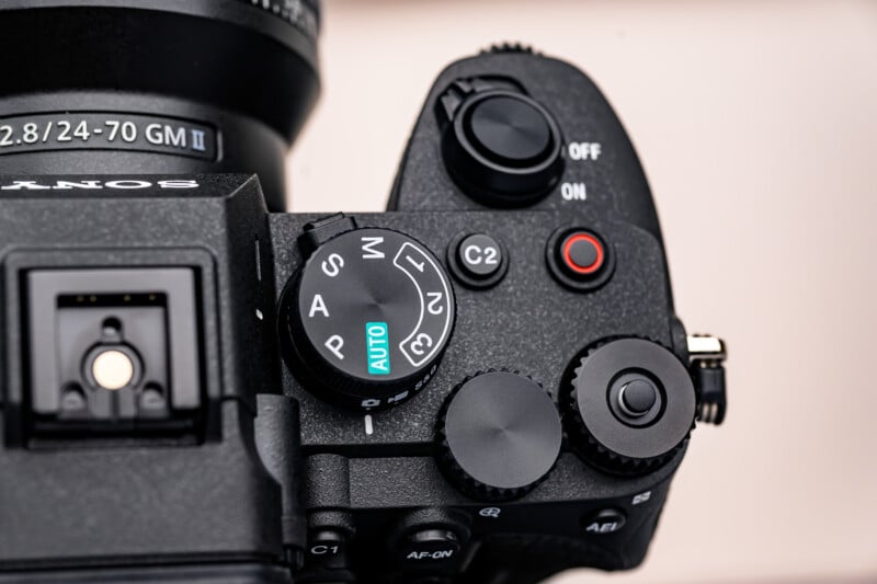 Close-up of a black Sony camera's top controls, showing mode dial with "AUTO" highlighted, power switch, shutter button, and various control buttons and dials. The lens information is partially visible at the top.