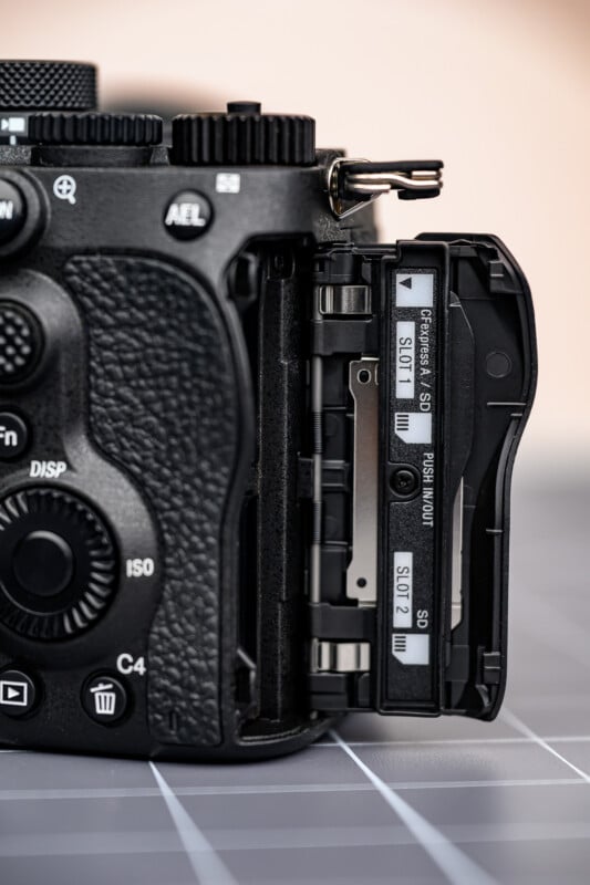 A close-up of a black digital camera with its side compartment open, revealing two SD card slots labeled “SLOT 1” and “SLOT 2” on a gray grid-patterned surface.