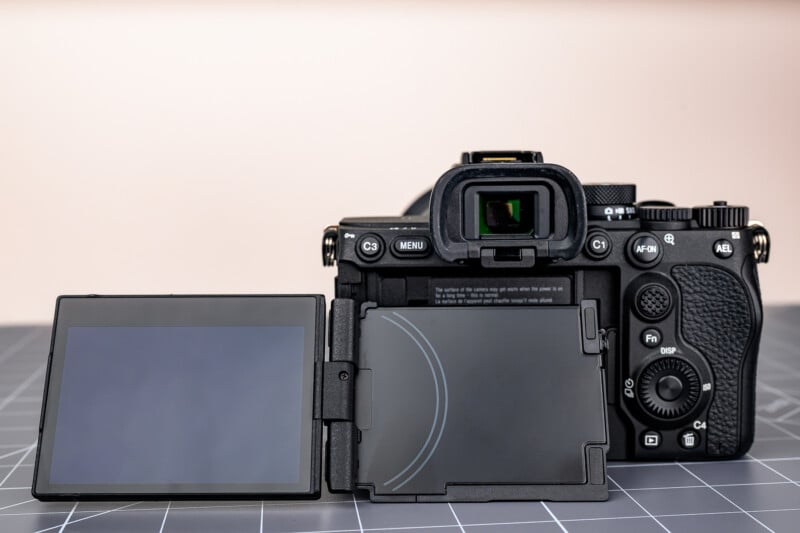 A digital camera with a fully articulated LCD screen is displayed on a grid-patterned surface. The screen is flipped out and angled to the left, showing the camera’s controls and viewfinder from the back.