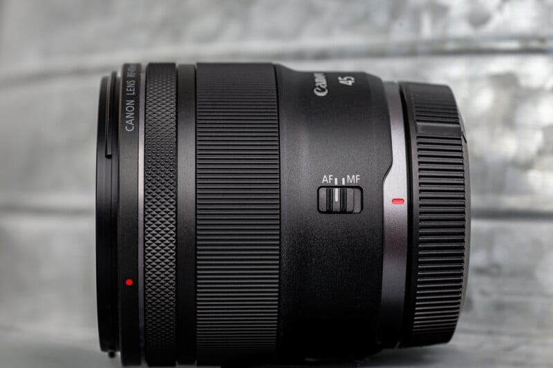 A close-up side view of a Canon RF 85mm camera lens, showing the lens body, focus ring, and autofocus/manual focus switch against a blurred metallic background.