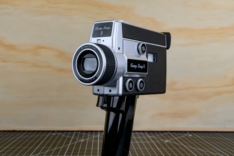 A vintage black and silver Camp Snap 8 handheld movie camera is displayed on a grid-patterned surface with a light wooden background.