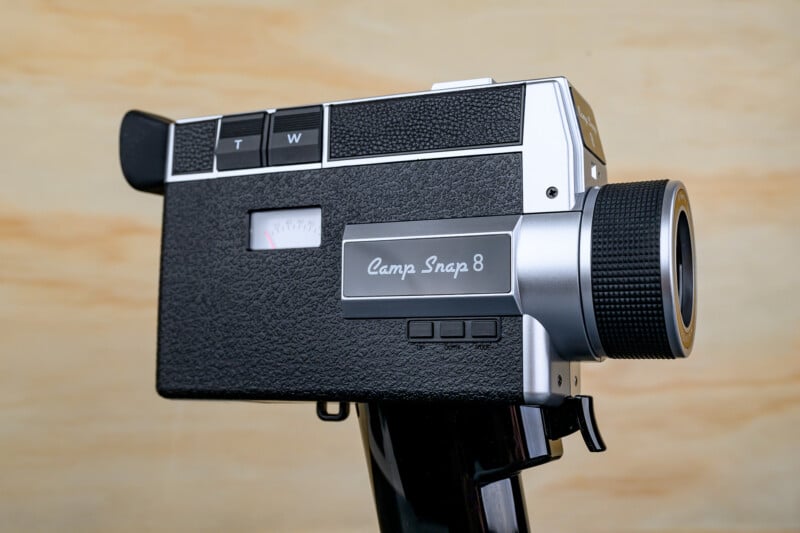 A vintage black and silver Camp Snap 8 movie camera with a textured body, lens, and control buttons, shown against a light wood background.