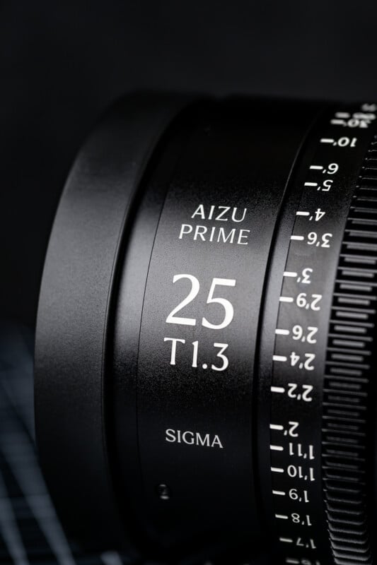 Close-up of a black Sigma Aizu Prime camera lens showing white text “25 T1.3” and focus distance markings. The image highlights the lens branding and precise details on a dark background.