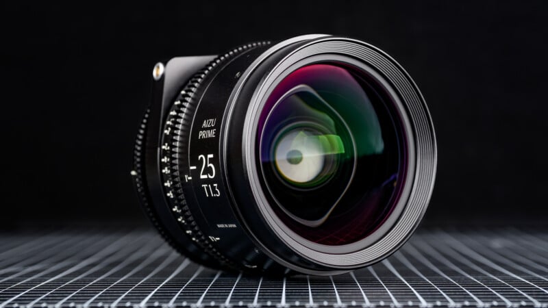 A close-up of a 25mm T1.3 cinema camera lens with a colorful reflection on its glass, placed on a grid-patterned surface against a dark background.