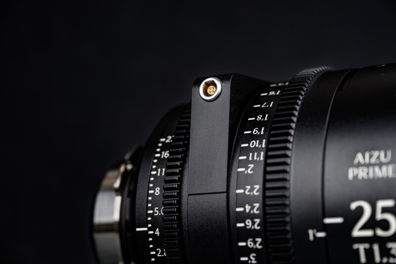 Close-up of a camera lens showing detailed focus and aperture markings, with a black metal exterior and a small gold connector on a bracket, set against a dark background.