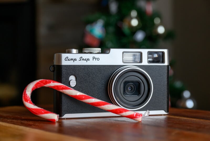 A vintage-style "Camp Snap Pro" camera sits on a wooden surface with a red and white candy cane in front of it; a blurred, decorated Christmas tree is visible in the background.