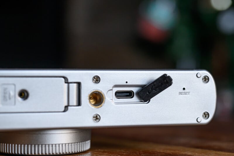 Close-up of an electronic device’s silver back panel showing a USB-C port with a black rubber cover, a reset button hole, a screw, and a metal fitting, all on a wooden surface.