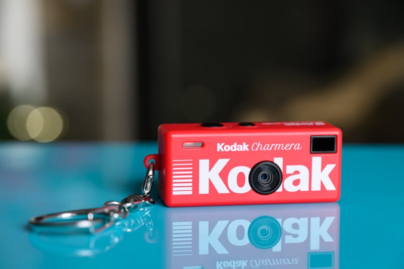 A small red Kodak camera keychain labeled "Kodak Charmera" sits on a shiny blue surface with a blurred dark background.
