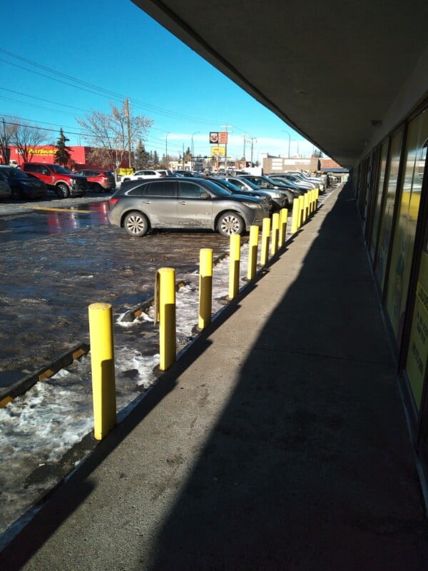 A row of yellow bollards lines a sidewalk beside a parking lot with cars on a sunny winter day; some ice and snow are visible along the edge of the lot.