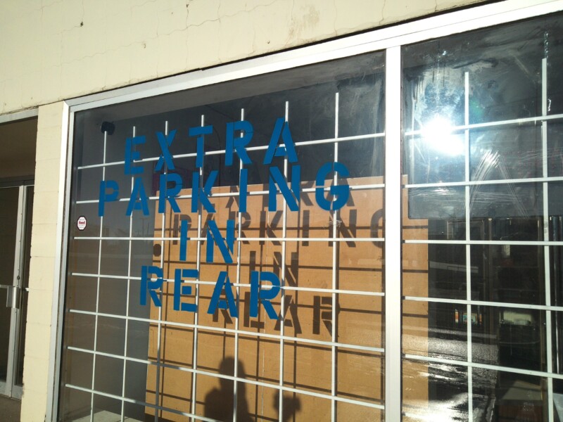 A glass storefront window with a metal grid and blue letters reading "EXTRA PARKING IN REAR," with sunlight reflecting off the glass and the text casting a shadow on cardboard inside.